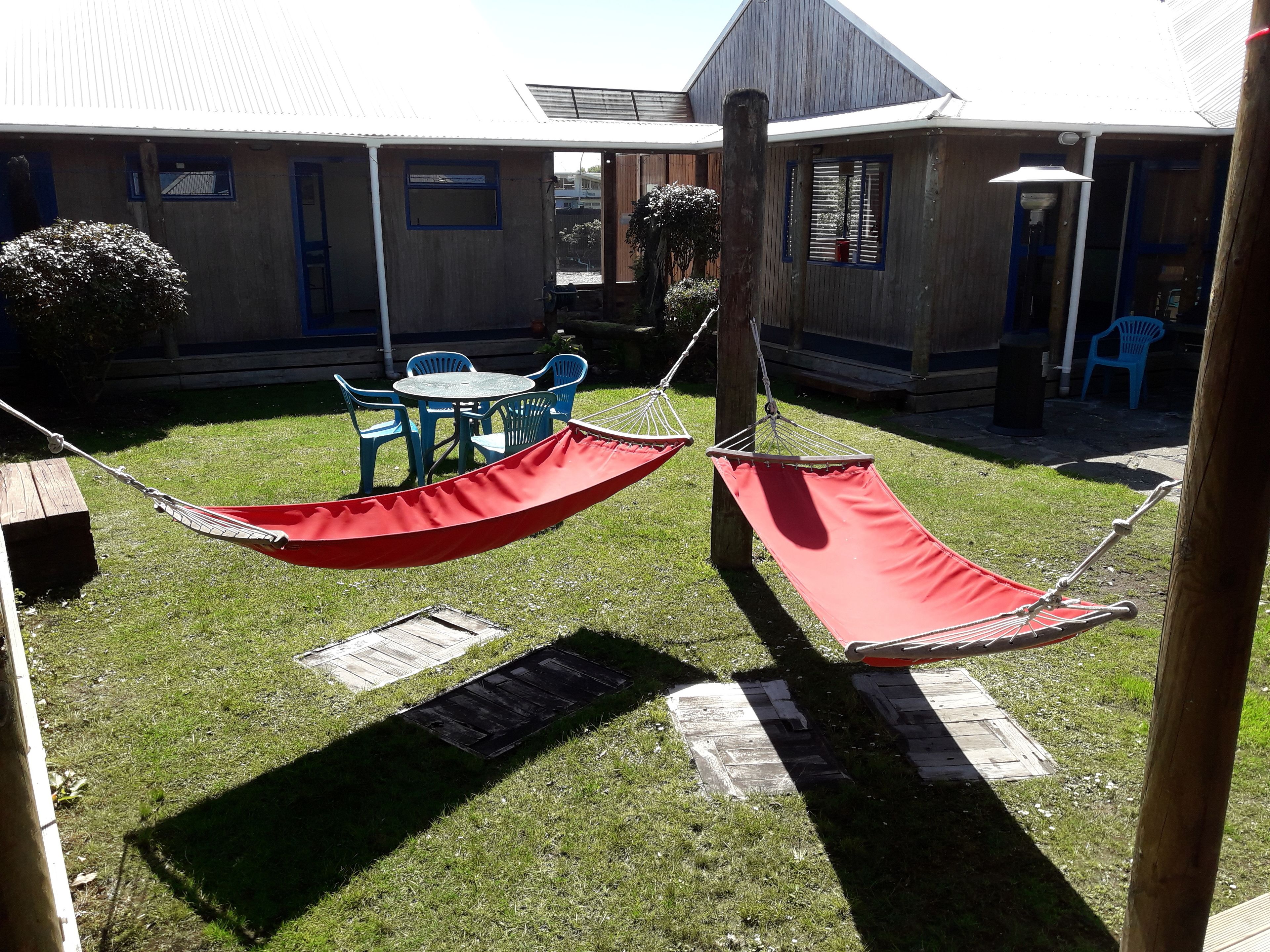 A backyard with two red hammocks, a wooden table with blue chairs, and a building with a blue door and windows.