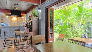 Un restaurante con mostrador de mármol, mesas y sillas de madera. Hay un televisor montado en la pared y un ventilador de techo. En el exterior, se ve una exuberante vegetación.