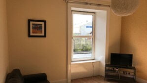 Un salon avec un canapé, un téléviseur posé sur un meuble, une fenêtre donnant sur une clôture et des bâtiments, et un cadre photo accroché au mur.