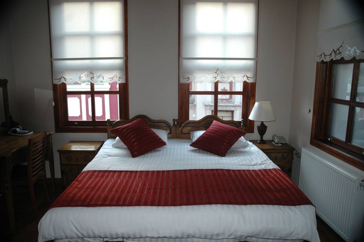 Een hotelkamer met een groot bed, twee rode kussens, een houten hoofdbord, een nachtkastje met een lamp en een raam met kantgordijnen.