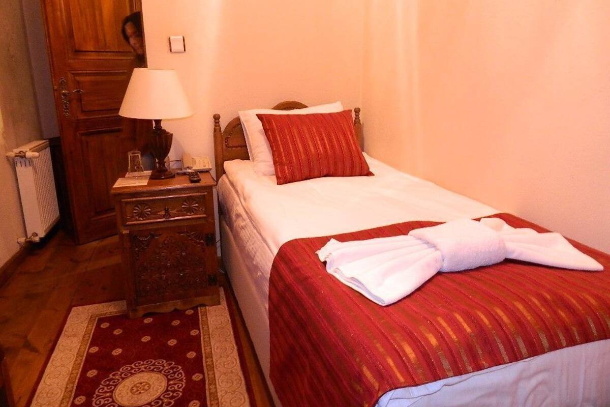 Een hotelkamer met een bed, een nachtkastje met een lamp, een houten garderobe en een rood vloerkleed met dessin.