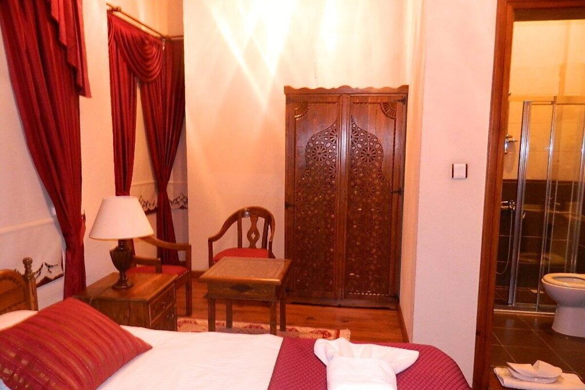 Hotelkamer met een bed, een houten garderobe, een stoel, een tafel met een lamp en een badkamer zichtbaar door een glazen deur.