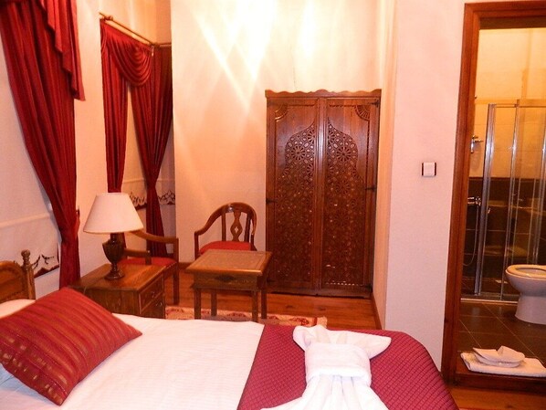 A hotel room with a bed, a wooden wardrobe, a chair, a table with a lamp, and a bathroom visible through a glass door.