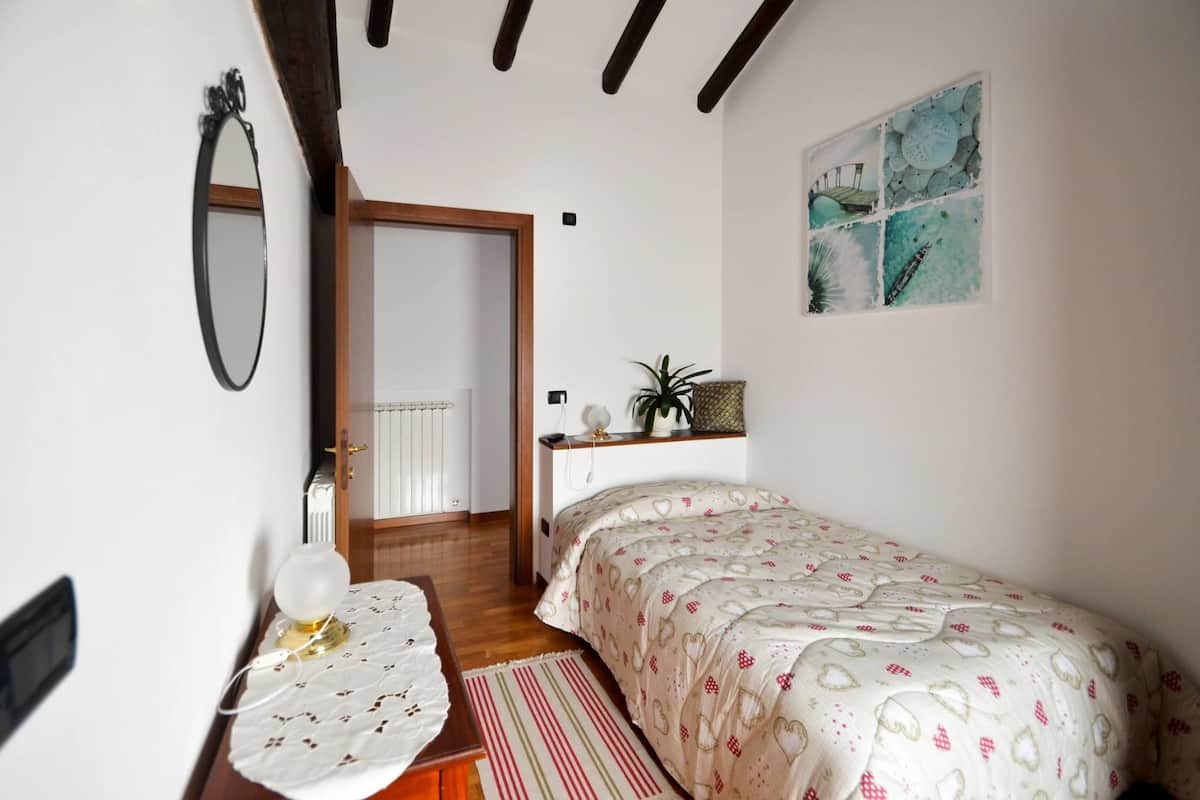 Een slaapkamer met een bed, nachtkastje, spiegel, radiator, deur en een muur met een schilderij.