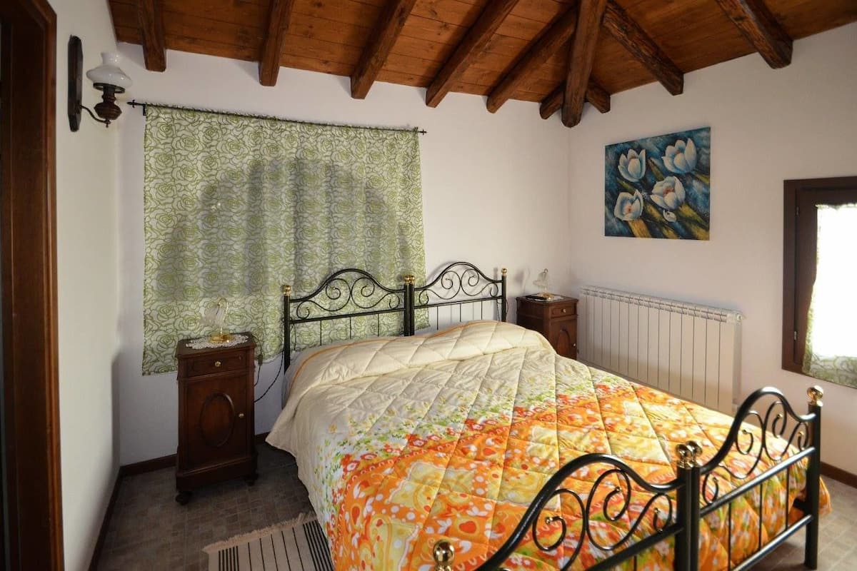 Een slaapkamer met een bed, houten hoofdeinde, nachtkastje, radiator, raam met een groene gordijn en een bloemrijk schilderij aan de muur.