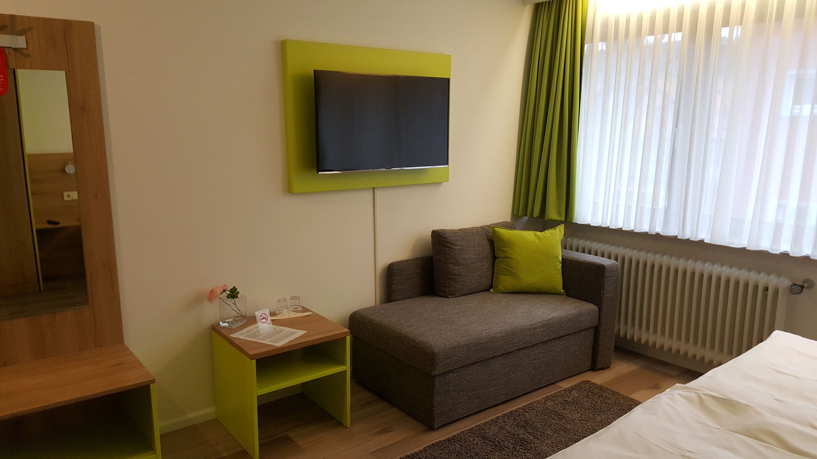 Una habitación de hotel moderna con un sofá gris, una mesita de madera pequeña, un televisor de pantalla plana montado sobre un panel verde, un radiador y una ventana con cortinas blancas traslúcidas.