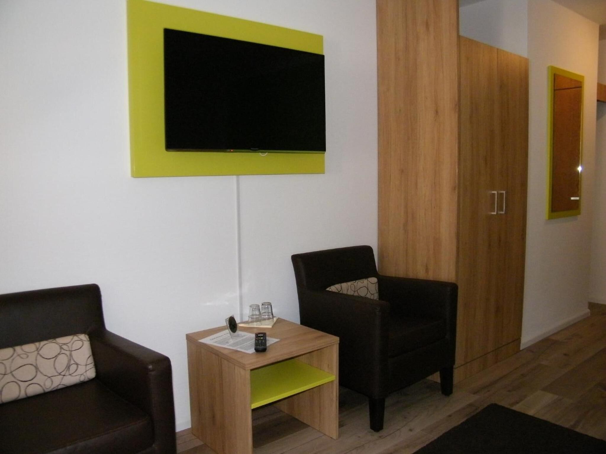 A modern living room with a flat-screen TV mounted on a white wall, two dark armchairs, a wooden side table with a remote control, and a wooden wardrobe.