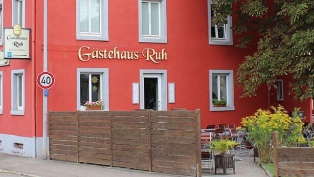 A red building with the sign "Gasthaus Ruh" indicating a guesthouse. There is a wooden gate, outdoor seating, and a tree in the background. - Gästehaus Ruh (Freiburg im Breisgau)