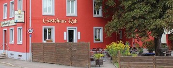 Un edificio rojo con el letrero "Gasthaus Ruh" que indica una posada. Se ve una verja de madera, mesas al aire libre y un árbol en el fondo.
