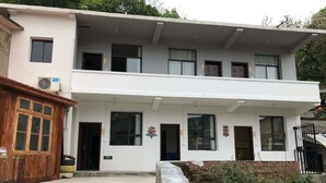 Un edificio de dos pisos con varias ventanas, un balcón y un pequeño espacio exterior con un muro de concreto y vegetación.