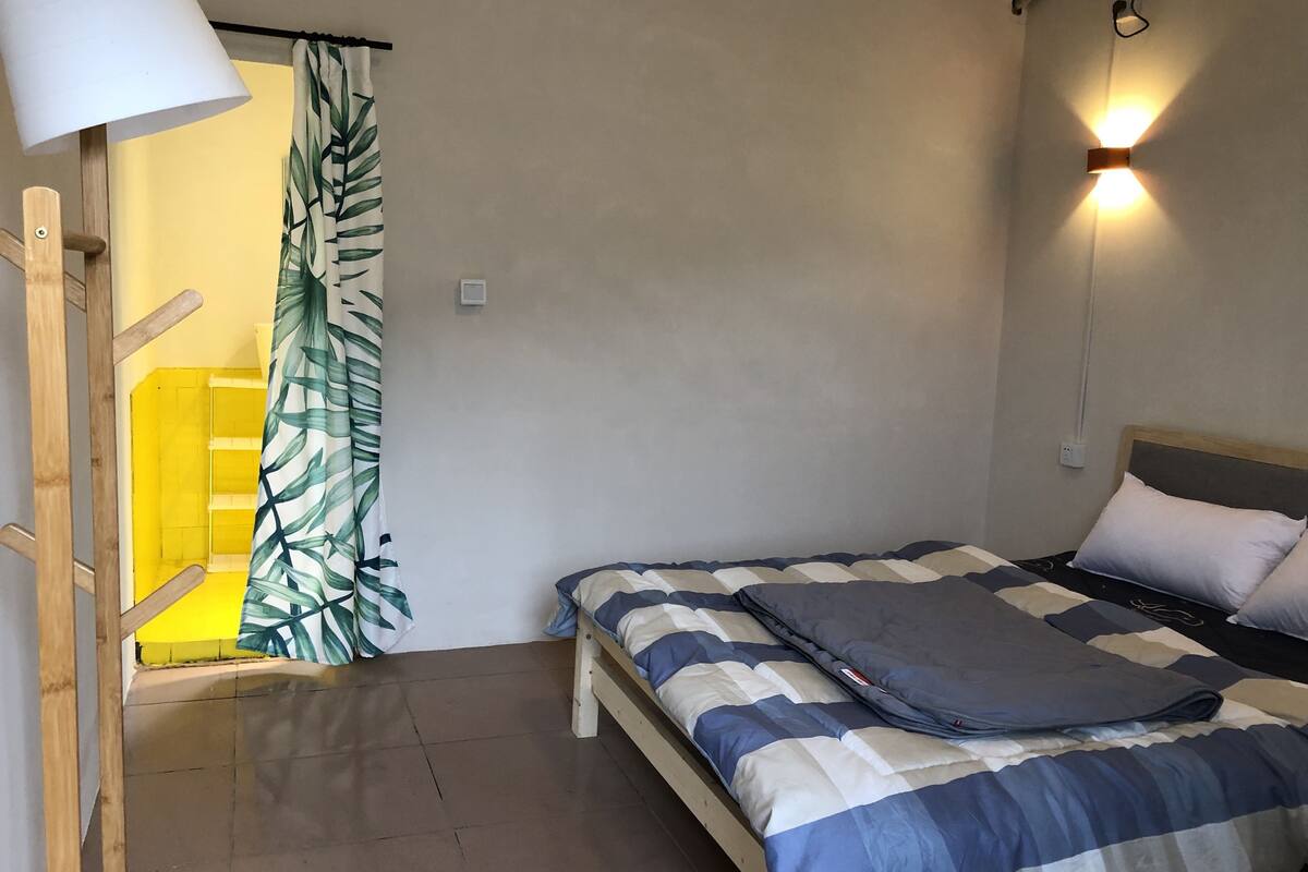 Quarto com cama, mesa de cabeceira, luminária, luz na parede e banheiro visível por uma cortina.