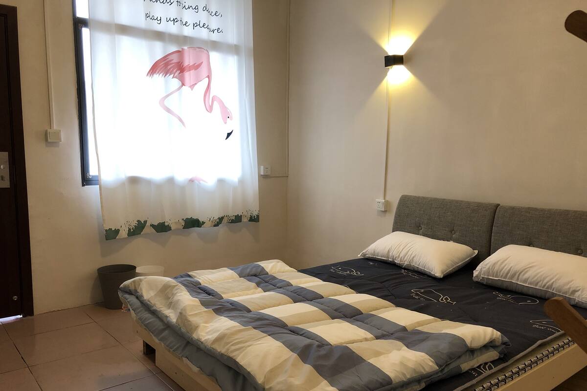 Quarto com cama, almofadas, luminária na parede e janela com cortina.