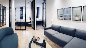 Un salón moderno con un sofá gris, una mesa de centro de vidrio y un sillón azul. Hay cuadros enmarcados en la pared y un piso de madera.