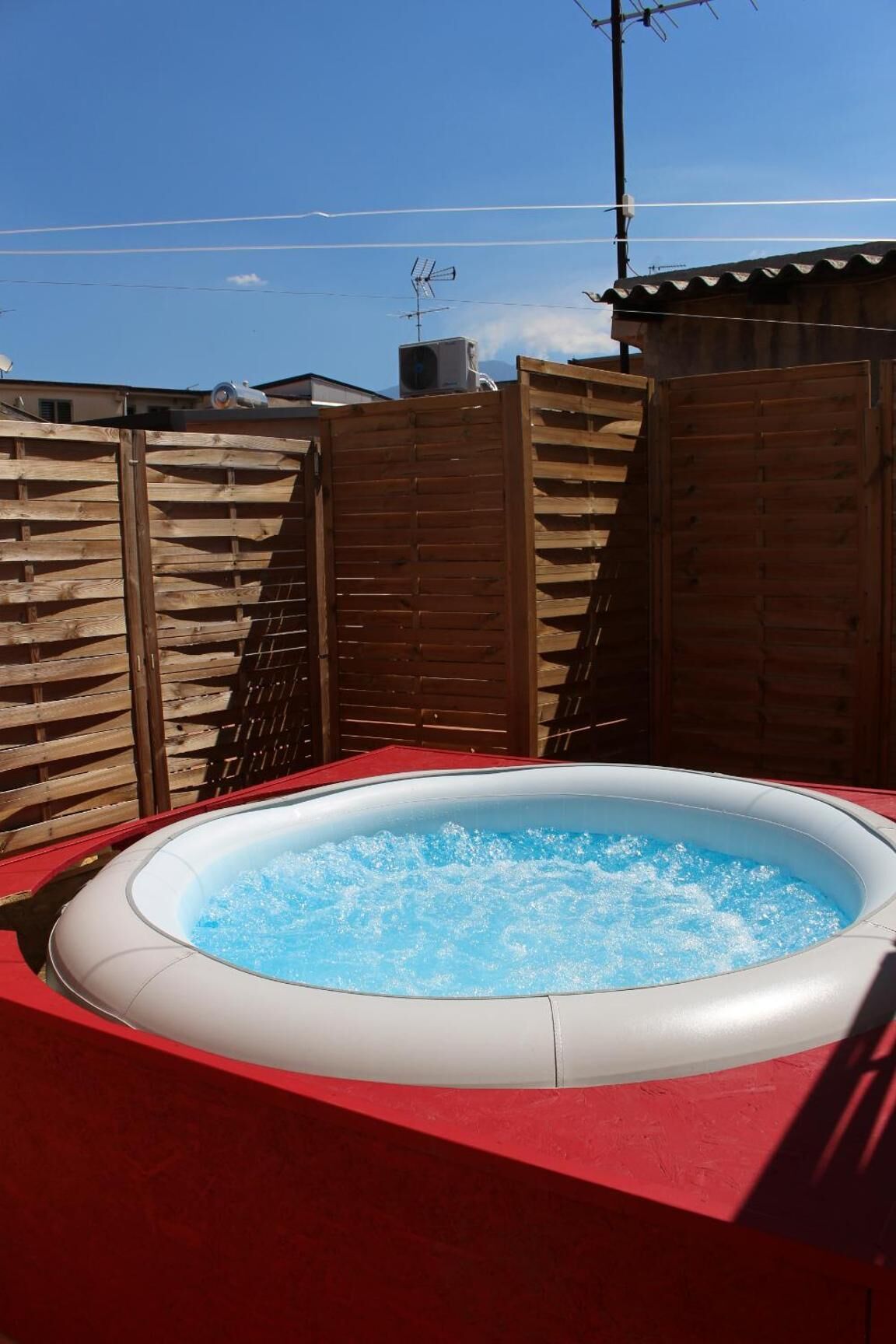 An outdoor hot tub with a red deck, surrounded by wooden fences, under a clear blue sky.