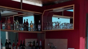 Uma área de bar com design em vermelho e madeira, apresentando um balcão com garrafas e copos, e três banquetas.