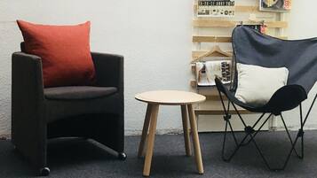 A room with a black armchair with a red cushion, a small round table, and a black folding chair. There are posters and magazines on a wooden rack against the wall.