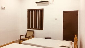 A room with two single beds, a wooden chair, a small table, a window with vertical blinds, a door, and an air conditioning unit. - Yarl Crown Inn (Kandy)
