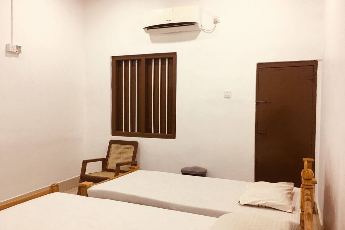 Quarto com duas camas de solteiro, uma cadeira de madeira, uma mesa pequena, uma janela com persianas verticais, uma porta e um aparelho de ar condicionado.