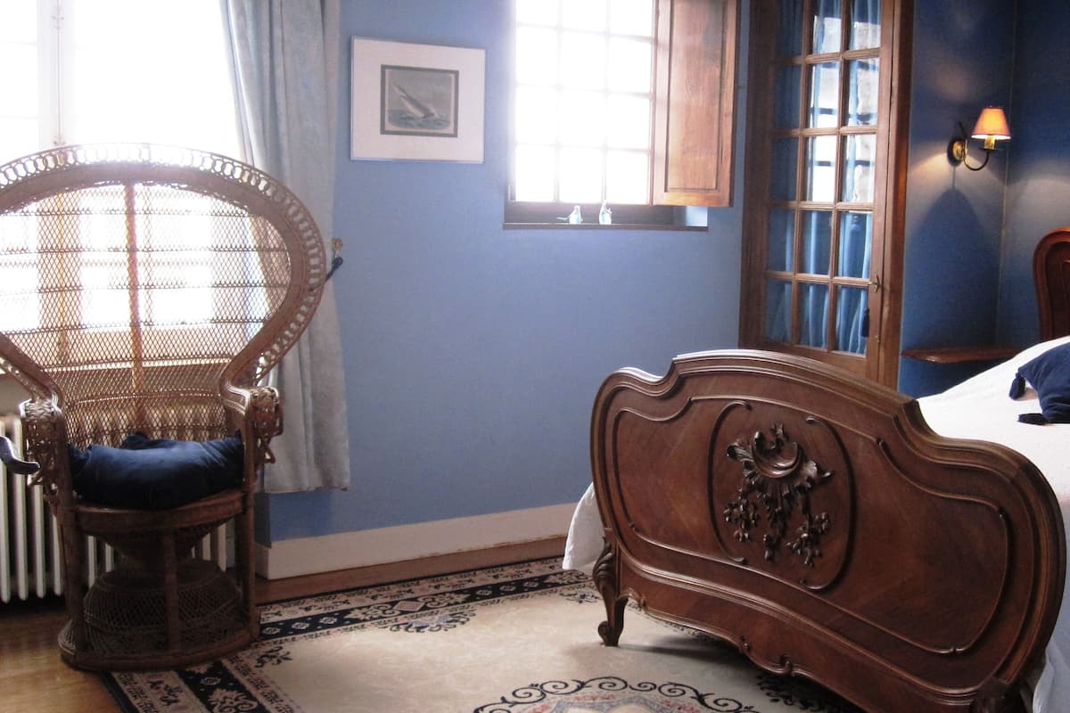 A bedroom with a wooden bed, a wicker chair, a window with curtains, and a wall-mounted lamp.