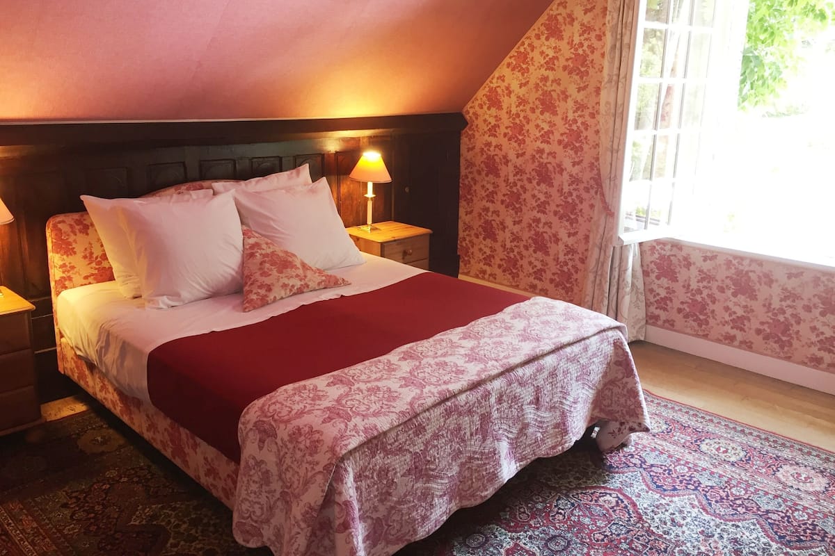 A bedroom with a bed, nightstands, a lamp, floral wallpaper, and a window with curtains.
