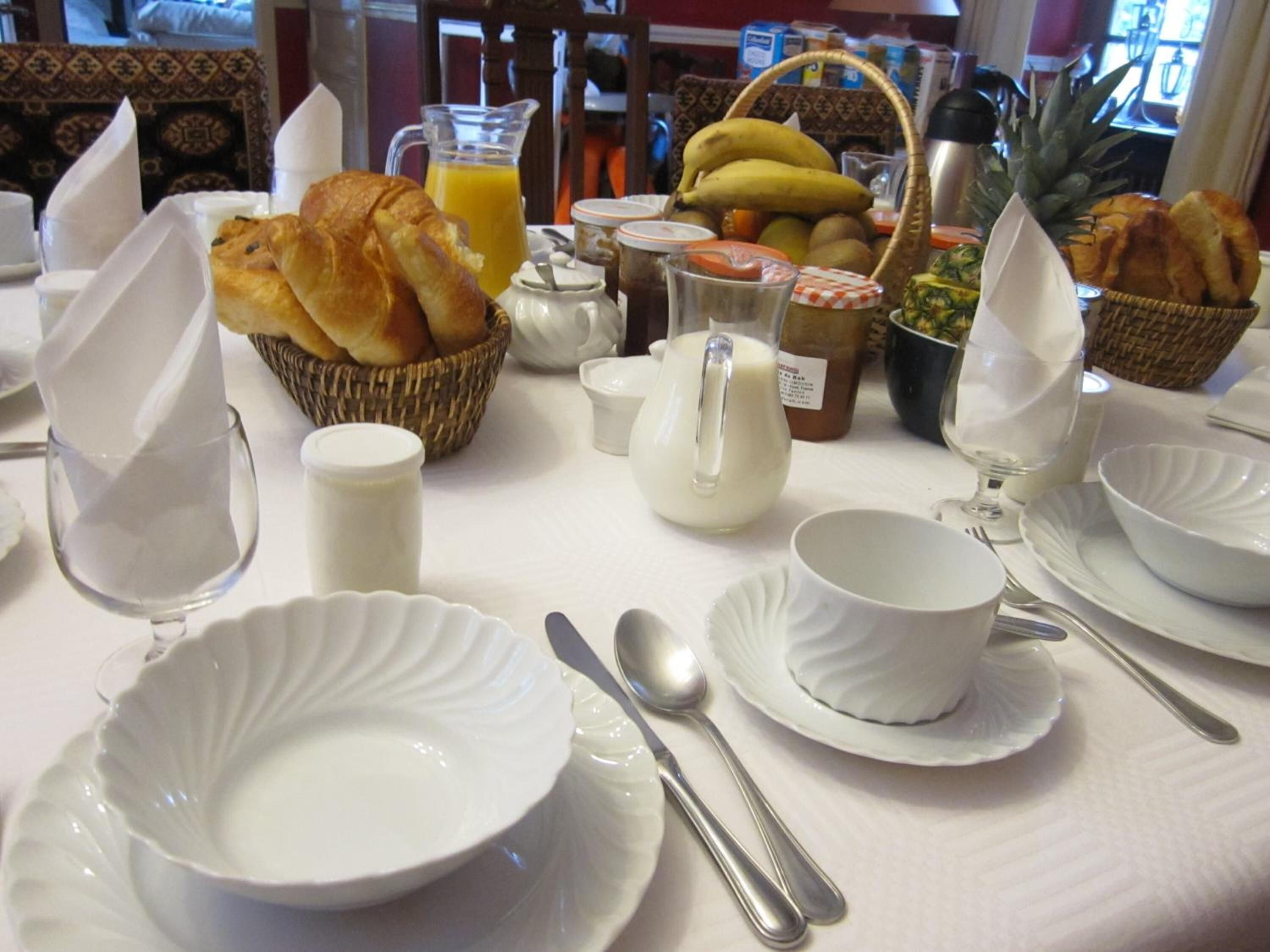 A breakfast table set with white dishes, bread, a pitcher of milk, a glass of orange juice, and a basket of fruits.