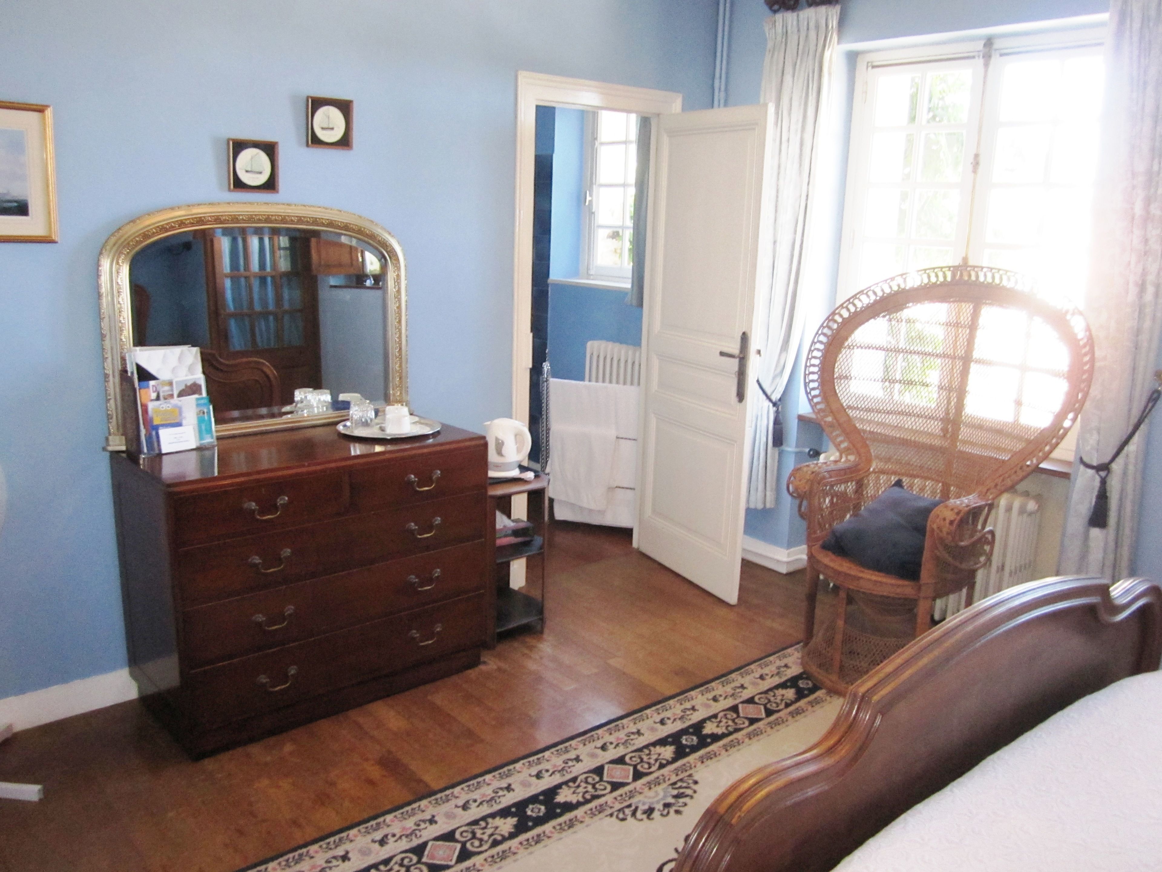 Une pièce comprenant une commode en bois, un miroir, une chaise en osier et une porte donnant sur une autre pièce.