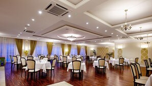 A spacious banquet hall with multiple round tables and chairs, a polished wooden floor, and elegant chandeliers.