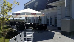 An outdoor seating area with tables and chairs under white umbrellas, a white chair, and a building with large windows and a stone pillar.
