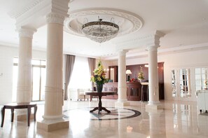 A spacious lobby with marble flooring, white columns, a central chandelier, and a reception desk.