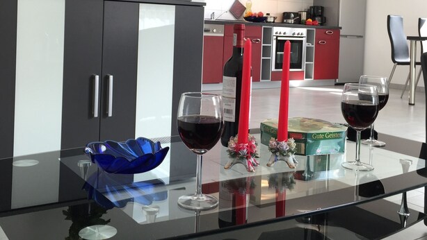 A modern kitchen with red and white cabinets, a black countertop, and a dining area with a glass table. - Bett & Bike Eden (Senftenberg)