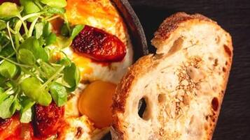 A dish of baked eggs with greens and tomatoes, accompanied by a slice of bread on a dark surface.