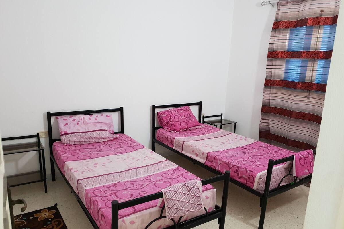 A room with two single beds, each with a black metal frame and pink bedding. There is a small table next to each bed, and a curtain with a striped pattern is partially visible.
