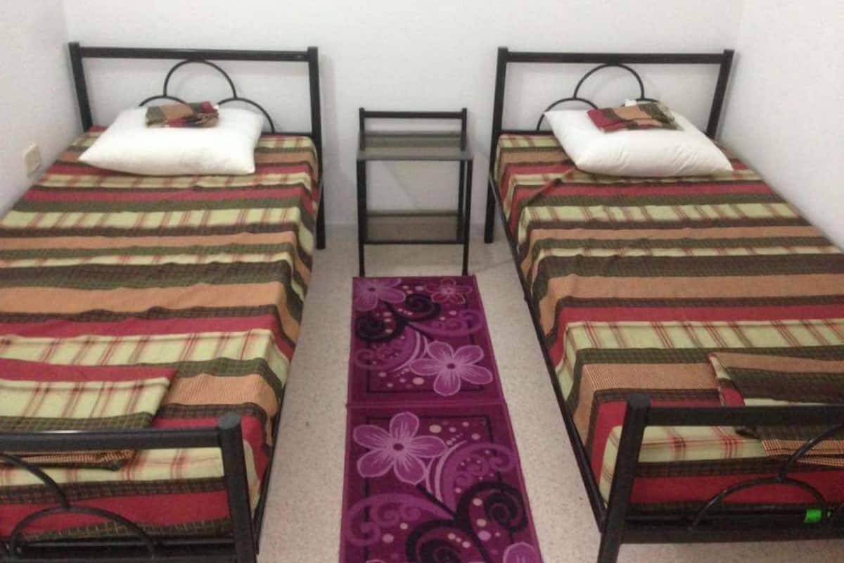 A room with two single beds, each with a striped bedspread and a pillow. A small table is placed between the beds, and a purple floral rug is on the floor.