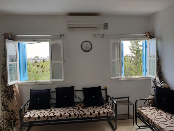 Quarto com duas janelas abertas, cama com roupa de cama estampada, uma mesa pequena e um aparelho de ar condicionado.