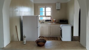 Een compacte keuken met witte kastjes, een koelkast, een magnetron en een spoelbak. Er is een raam met blauwe luiken en een klein vloerkleed.
