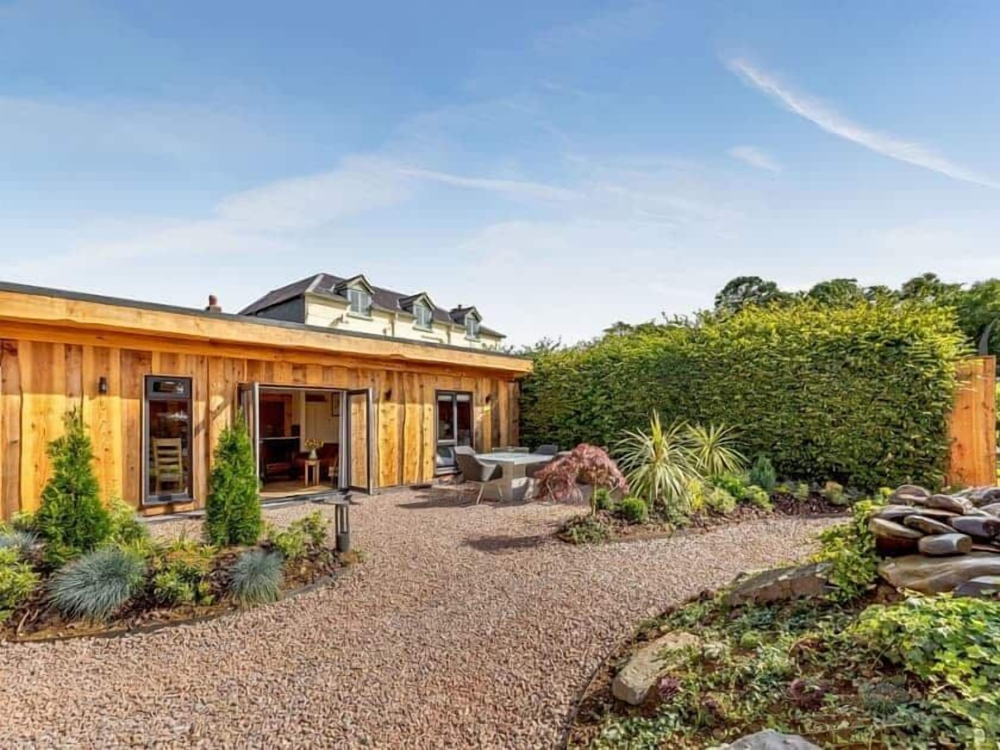 Una casa de madera con camino de grava, un patio con mobiliario exterior y un jardín bien cuidado con diversas plantas y un seto.