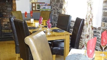 Un comedor con una mesa de madera dispuesta para cuatro personas, rodeada de sillas de cuero. Se observa una pared de piedra y una chimenea al fondo.
