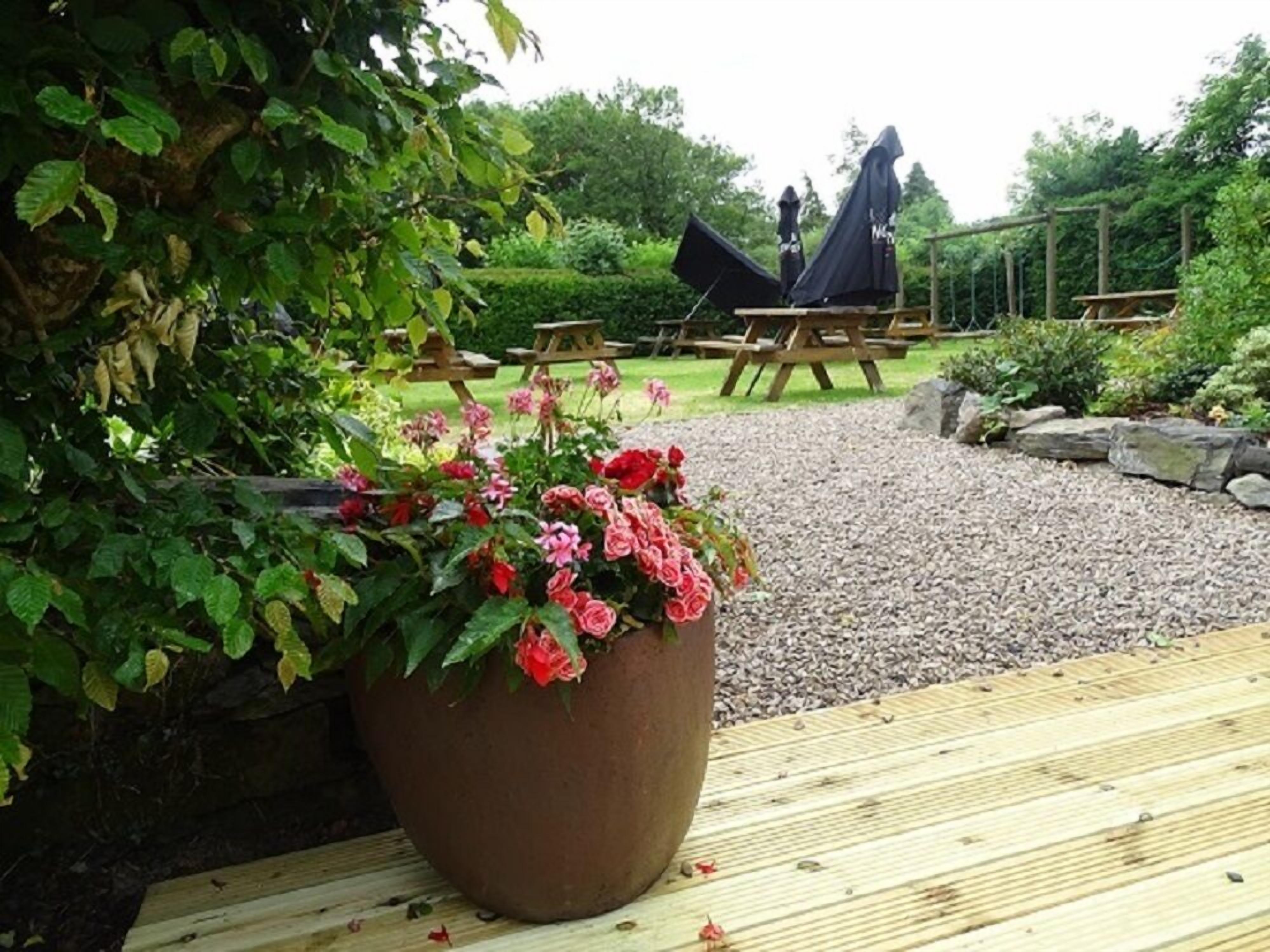 Un jardín con una maceta grande de flores rosas, mesas de picnic de madera y una estatua al fondo.