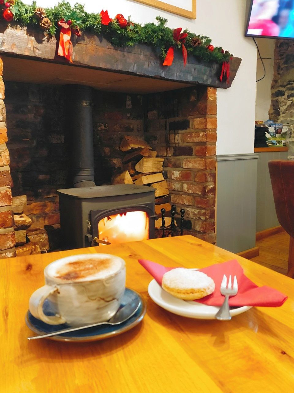 A cozy indoor setting with a brick fireplace, a table with a cup of coffee and a plate with a pastry, and a television mounted on the wall.