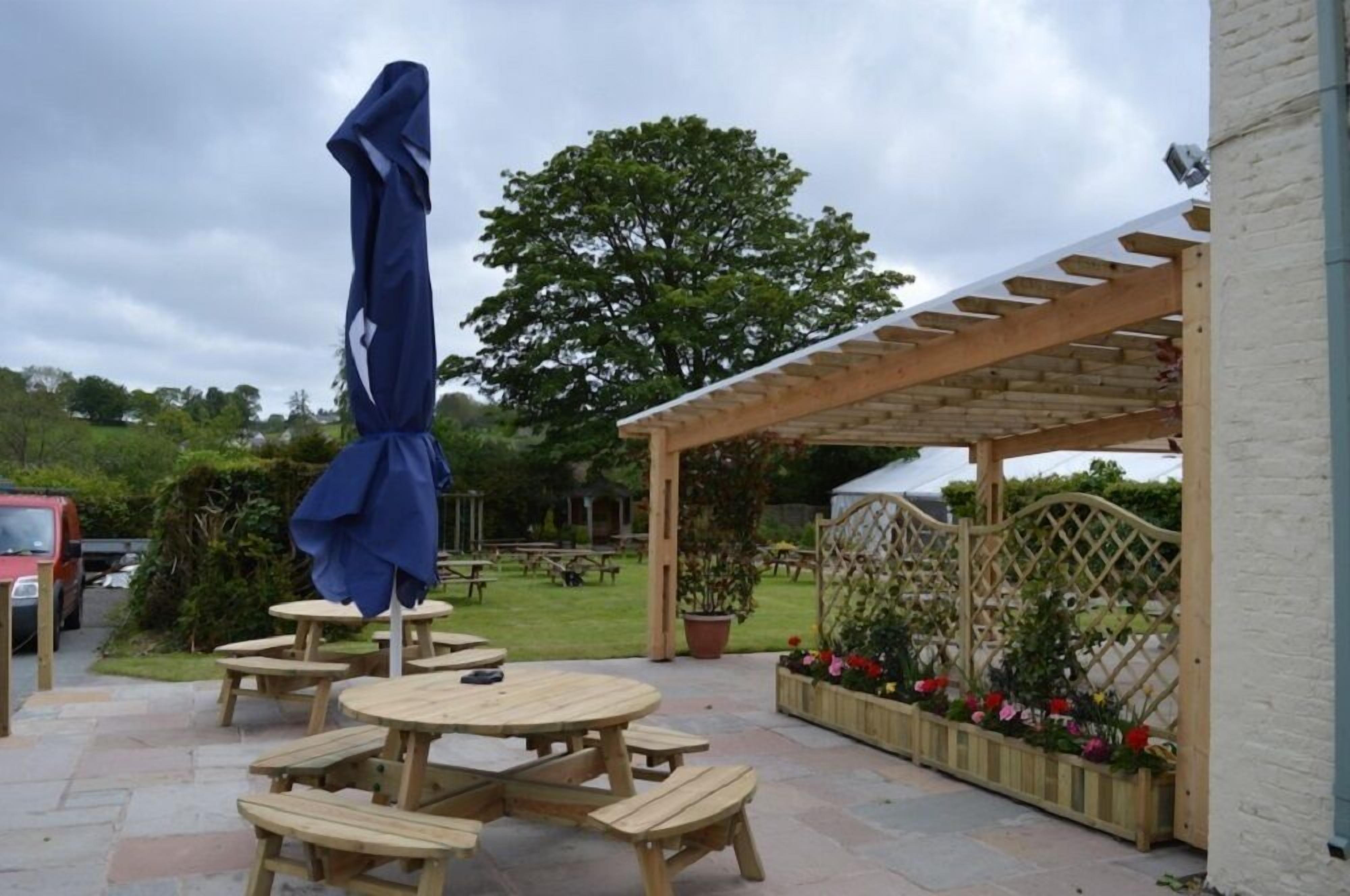 Une terrasse extérieure avec des tables et des bancs en bois, une pergola et un espace jardin agrémenté de fleurs et d’un arbre.