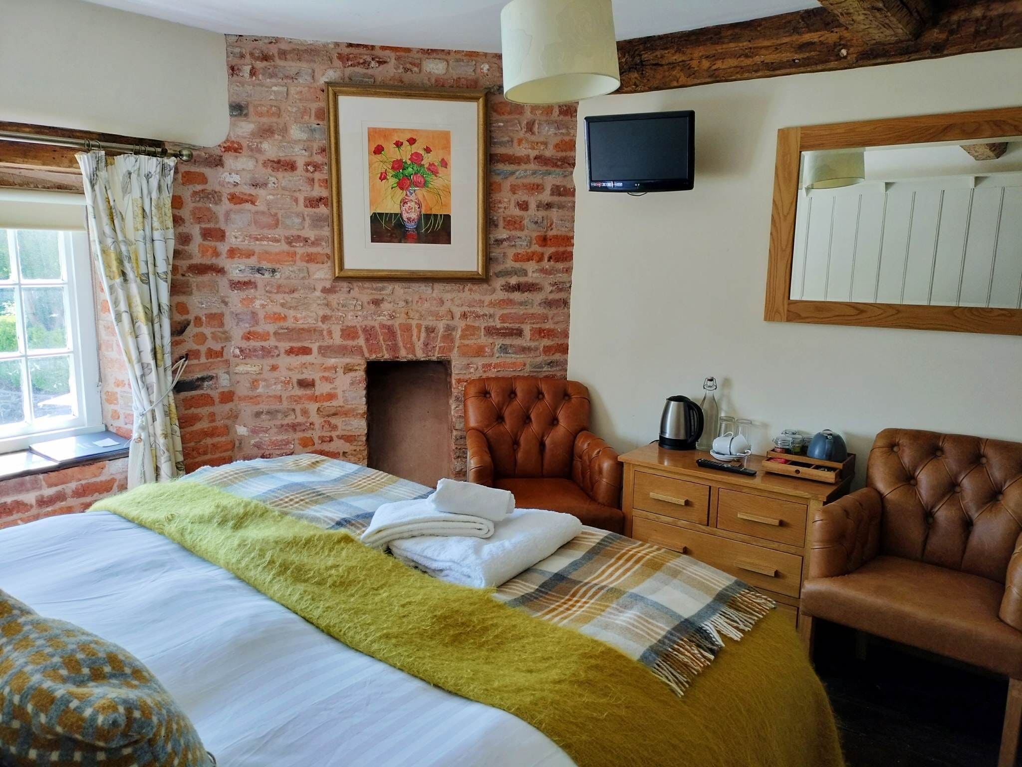 Una habitación acogedora con pared de ladrillo, chimenea, cama con manta escocesa, sillón de cuero y televisor en la pared.