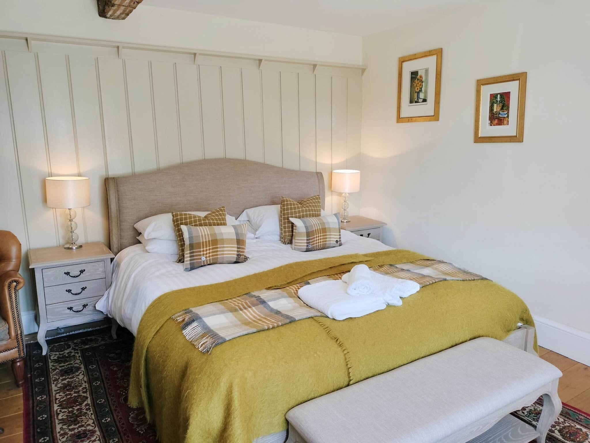 A neatly made bed with a plaid blanket, two bedside lamps, a bench, and a small table with a lamp.