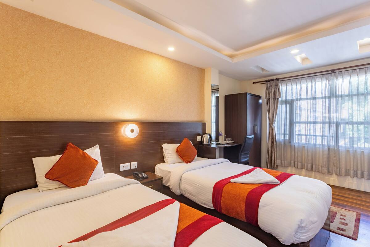 Ein Hotelzimmer mit zwei Betten, einem Nachttisch, einem Schreibtisch, einem Spiegel und einem Fenster mit Vorhängen.