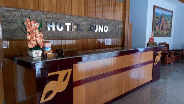 A hotel reception desk with a wooden finish, a sign that reads "HOTEL JUNO," and a reception area with a framed picture on the wall.
