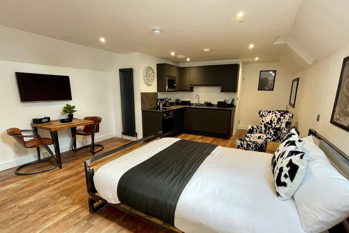A modern bedroom with a large bed, a desk, a chair, a sofa, and a kitchenette in the background.