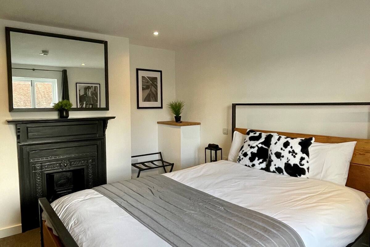 A bedroom with a large bed, a wooden headboard, a fireplace, a mirror, and a small table with a plant.