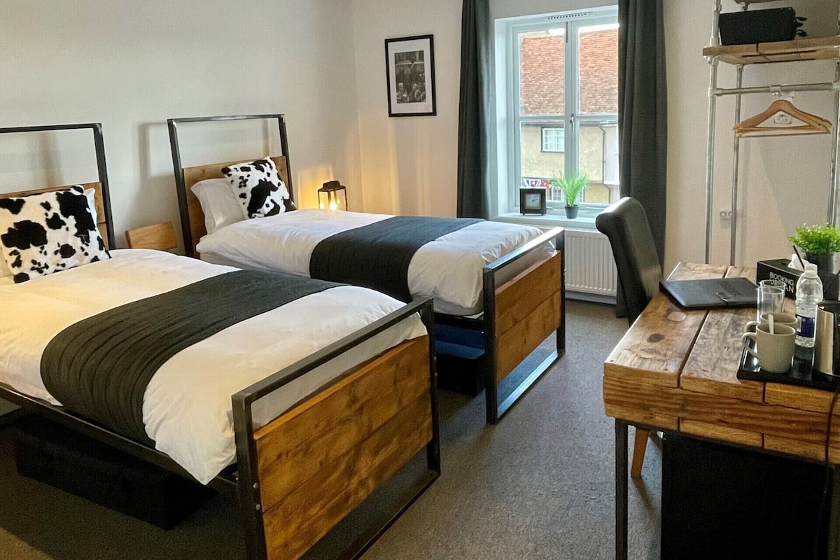 A hotel room with two beds, a desk, a chair, a window with curtains, a radiator, and a framed picture on the wall.