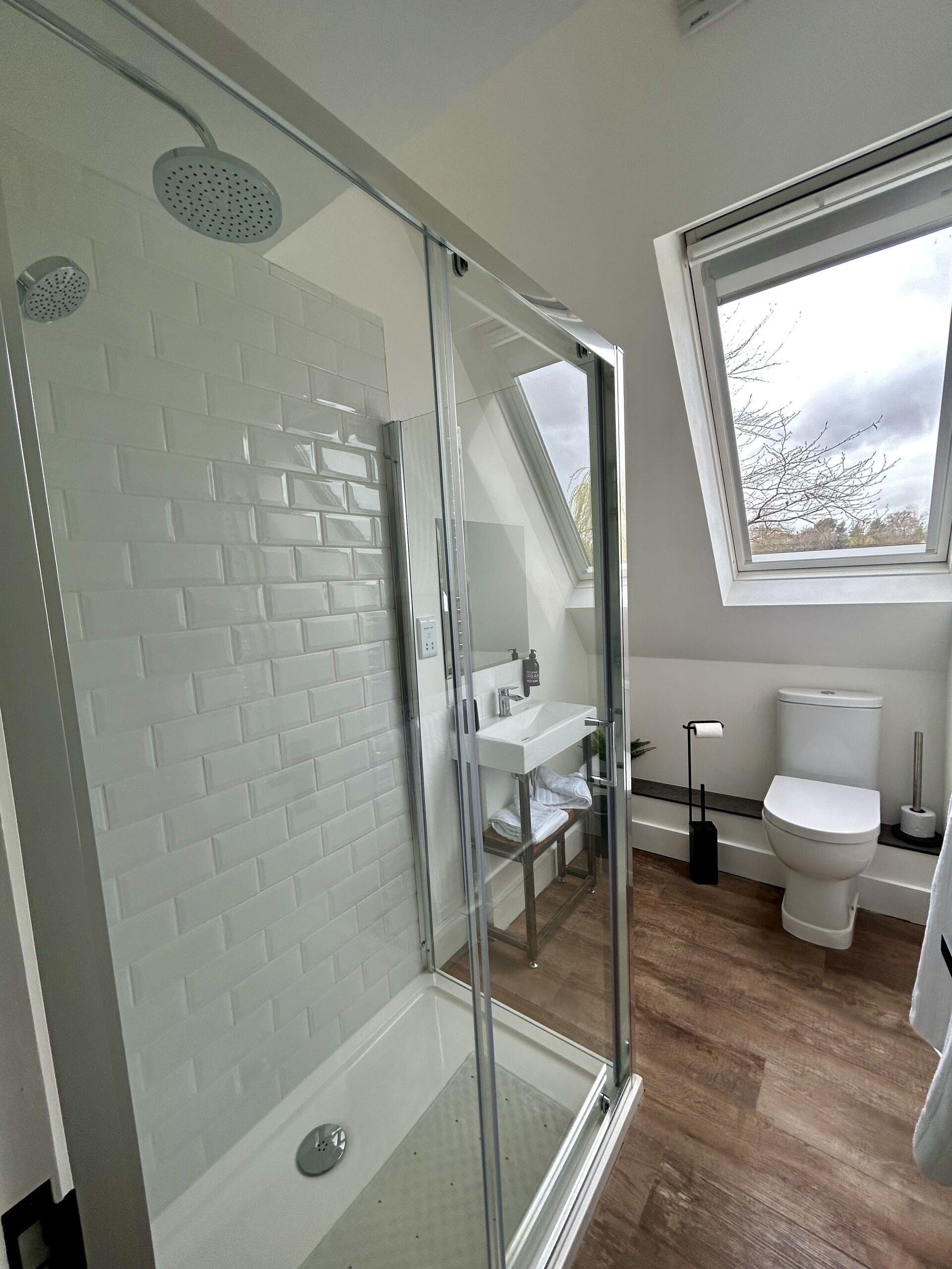 Un baño moderno con cabina de ducha de vidrio, paredes con azulejos blancos, suelo de madera, tragaluz y un inodoro blanco.