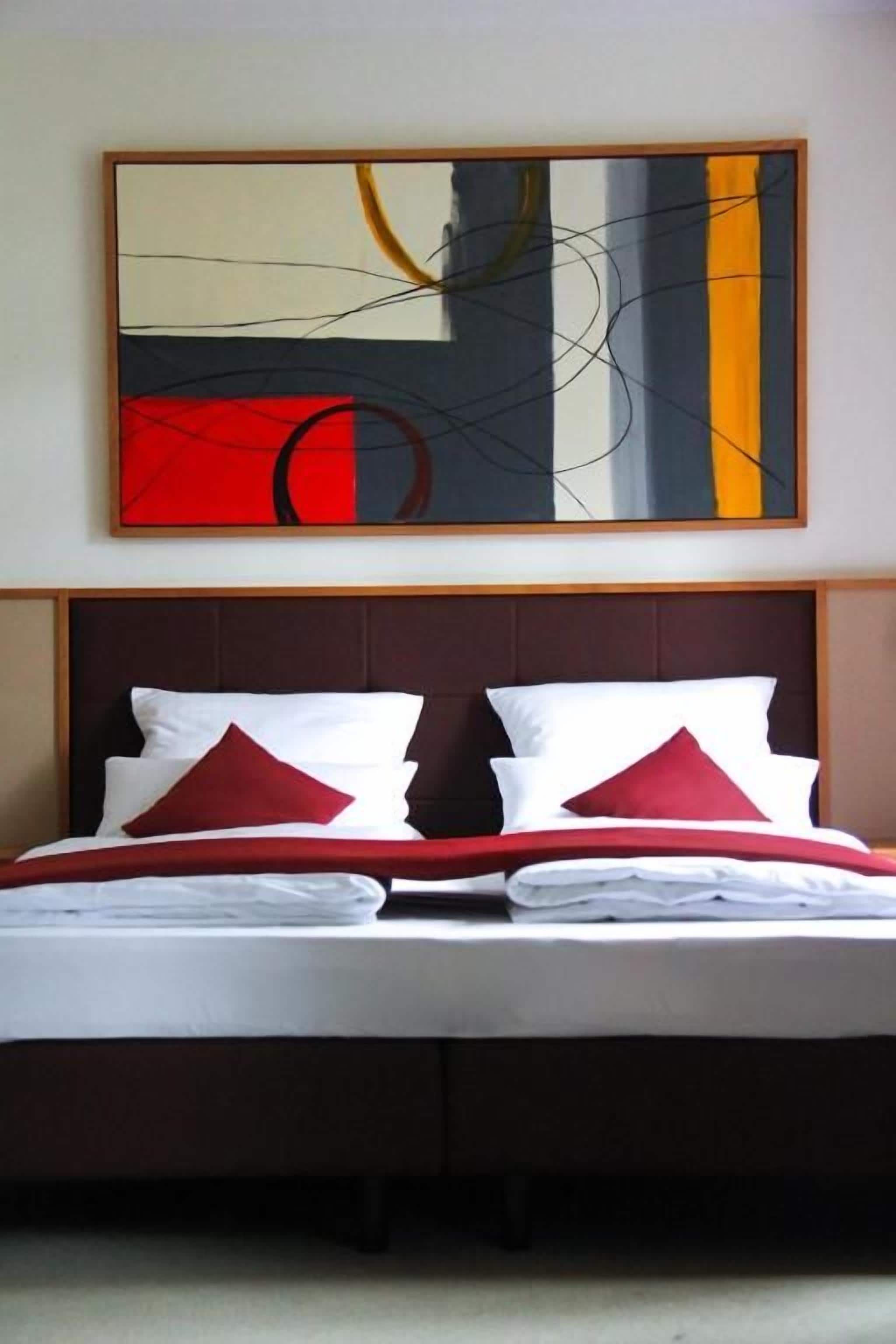 A bed with white linens and red pillows, a brown headboard, and a large abstract painting above it.