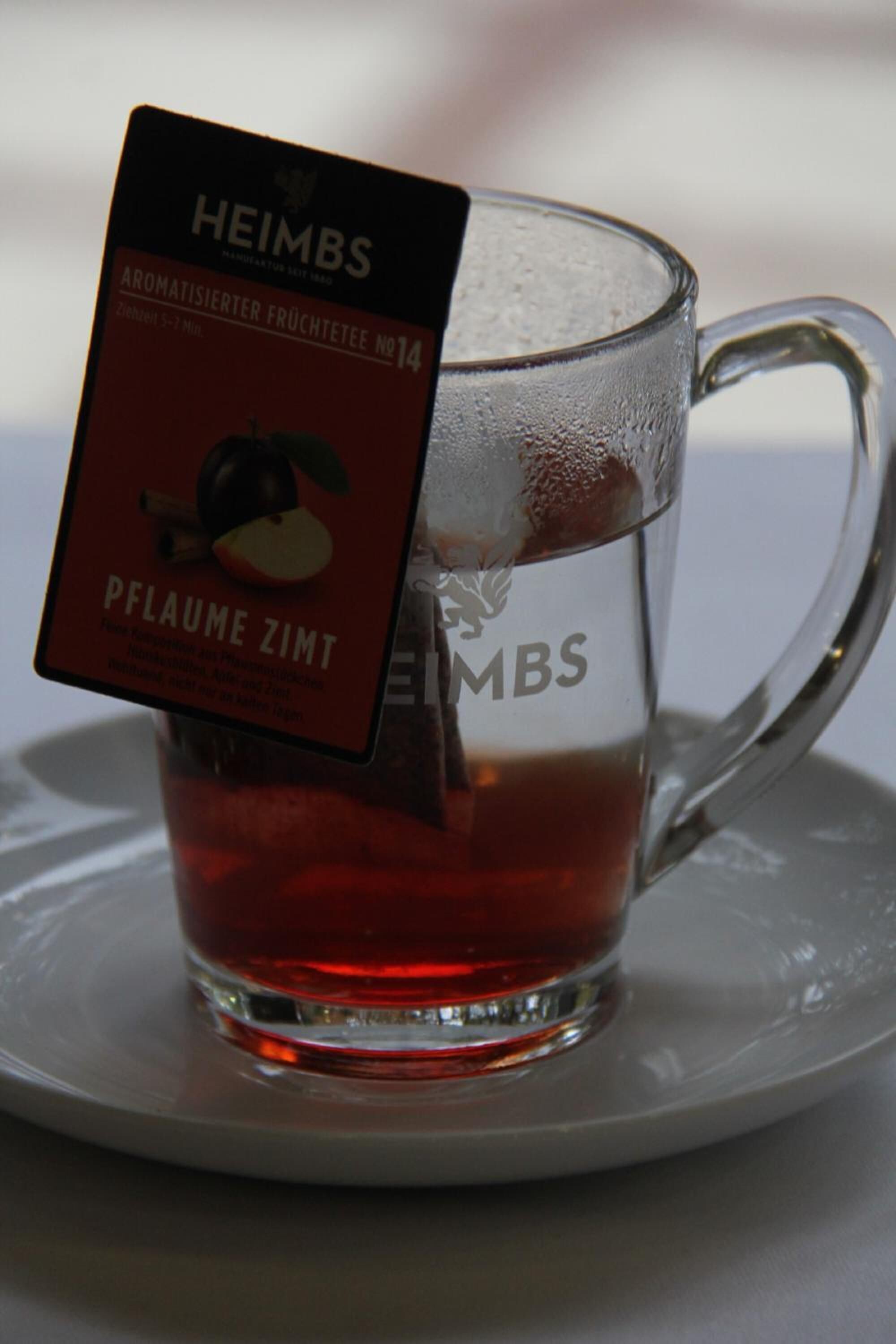Une tasse en verre transparent contenant un sachet de thé étiqueté « HEIMBS » et « PFLAUME ZIMT », posée sur une soucoupe.